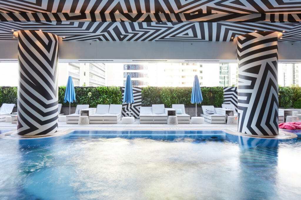 A green wall in our outside pool area at the W Hotel, Brisbane. In front of the green wall is three closed blue umbrellas, pool seating, and a well-maintained blue pool. Featuring striking black and white striped pillars and a ceiling. Maintained by Advance Plants